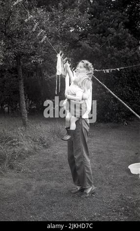 1960s, historical, helping mummy put washing on the clothes line, 'I will do the socks!'....outside in a back garden, a mother holding up her little girl, putting clothes on the line to dry, England, UK. Stock Photo