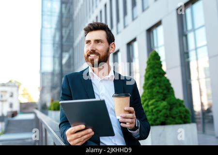 Cheerful confident young caucasian man with beard and tablet, looks at free space on balcony of office building Stock Photo