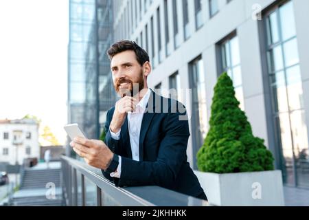 Smiling young european businessman with beard in suit thinking, typing on phone, looking at empty space Stock Photo