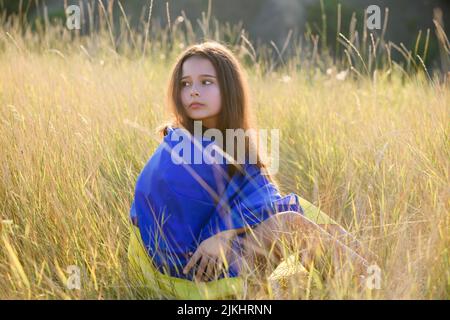 A teenage girl sits in tall wild grass on the slope of a ravine with a Ukrainian flag on her shoulders. Stock Photo