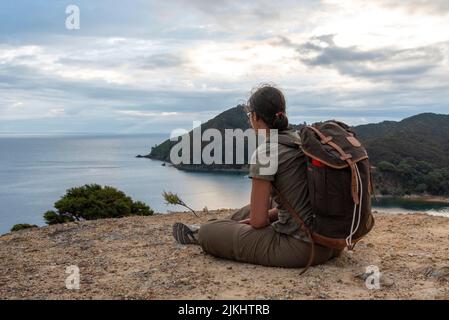 Woman sitting on a rock enjoying the magnificent view of Blind Bay on Great Barrier Island, New Zealand Stock Photo