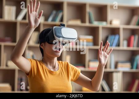 Amazed excited young asian girl in VR glasses raises hands, exploring virtual reality world at home alone Stock Photo