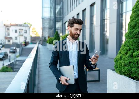 Smiling confident young businessman with beard in suit with laptop typing on smartphone Stock Photo