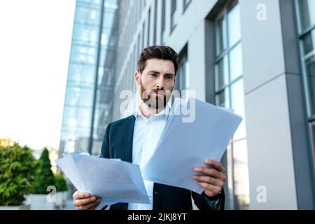 Busy confident young businessman with beard in suit works with documents near office building Stock Photo