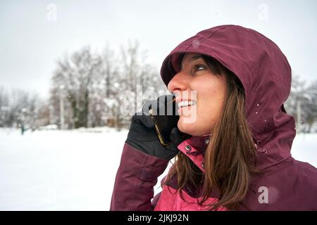 A closeup shot of a Caucasian female with a hoodie talking to phone with snow ground and trees around