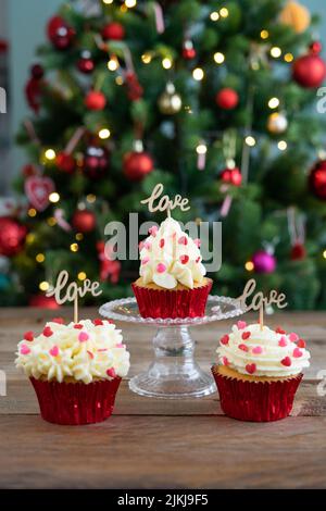 Three cupcakes decorated on wooden background with LOVE sign and out-of-focus Christmas tree in background. Copy space. Vertical photo. Stock Photo