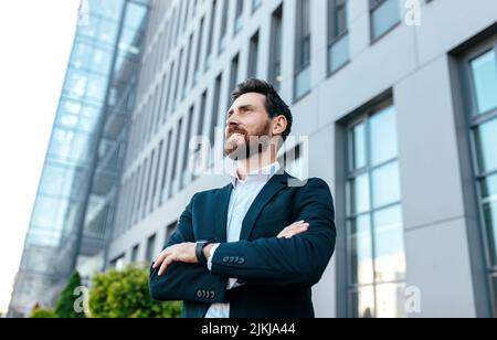 Smiling confident handsome young businessman with beard in suit with crossed arms on his chest looks at empty space Stock Photo