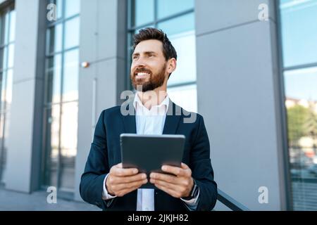 Happy confident millennial european man with beard in suit use tablet, look at empty space near modern office Stock Photo