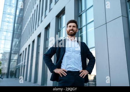Cheerful confident handsome millennial businessman with beard in suit stands near modern office building Stock Photo