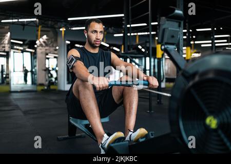 Sporty Young Black Man Exercising With Rowing Machine At Modern Gym Stock Photo