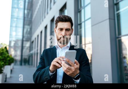 Serious confident millennial caucasian businessman with beard in suit typing on smartphone near office building Stock Photo