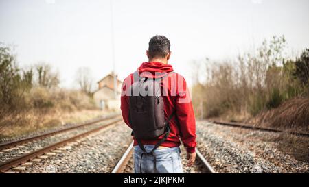 A shot of a man from his back with red hoody and backpack standing on a railroad Stock Photo