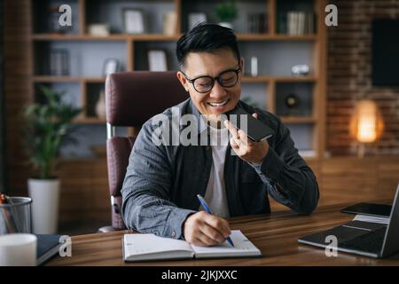 Smiling mature asian businessman in glasses talking on smartphone and making notes at workplace in home Stock Photo