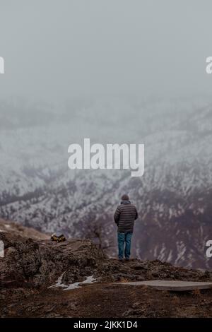 A vertical shot of a man standing on a cliff enjoying the beautiful snowy landscape with a toy car on the ground Stock Photo