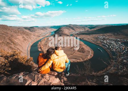 A back view of a couple sitting on the edge of a cliff looking at the river Stock Photo
