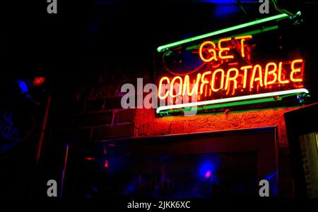 A view of a neon sign at a dark bar saying 'Get Comfortable' hanging on a brick wall Stock Photo