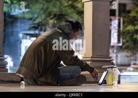 A young man sitting on the street and watching a video on a phone at night Stock Photo