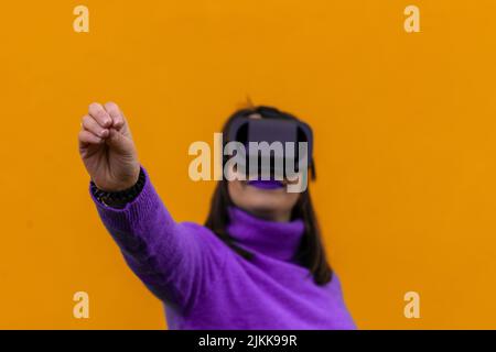 portrait of a smiling woman wearing virtual reality goggles with the gesture of picking up something imaginary with her hand on a yellow background. s Stock Photo