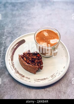 A vertical closeup of a cup of coffee with milk and a bitten slice of a pecan pie on the white plate Stock Photo