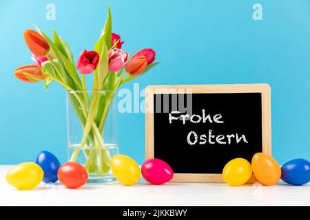 colorful tulips with chalkboard and easter eggs on a blue background with german text frohe ostern, in english happy easter Stock Photo