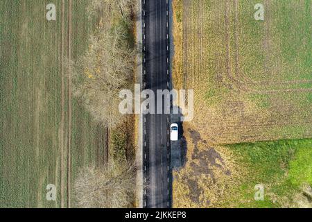 An aerial view of a white van parked on an asphalt roadside going through green fields with leafless trees on a sunny day Stock Photo