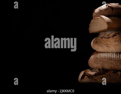 Heap of freshly baked loaves of bread with crust stacked on each other against black background in studio with obscure light Stock Photo