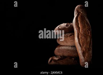 Heap of assorted brown freshly baked loaves of bread and baguette stacked on black background in studio with dim light Stock Photo