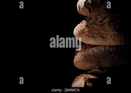 Heap of fresh baked loaves with crispy crust stacked on top of each other against black background in studio with dim light Stock Photo