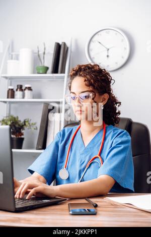 Young latin woman doctor in white medical uniform, with stethoscope using a computer laptop. Stock Photo