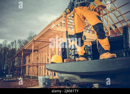 Contractor Worker and the Construction Site. Men with Large Hammer Staying on a Pickup Cargo Bed in Front of Wooden House Construction. Stock Photo
