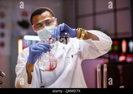 Science, chemistry, technology, biology and people concept - young african american male in protective mask and goggles scientist with test tube and microscope making research in clinical laboratory. Stock Photo