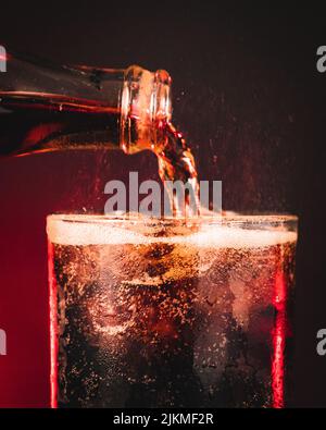 A vertical closeup of a bottle pouring soda into a with ice cubes in it against a red neon background Stock Photo