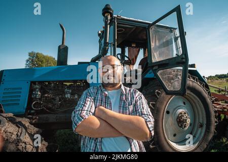 Portrait of satisfied tractor driver after work on agricultural field stands next to tractor. Farming and harvesting. Stock Photo