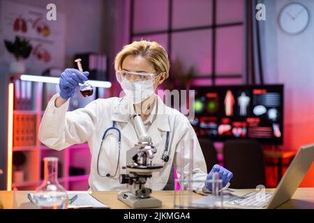Medical research laboratory: portrait of a attractive female scientist using digital laptop computer, analysing liquid biochemicals in a laboratory flask. Advanced scientific biotechnology laboratory. Stock Photo