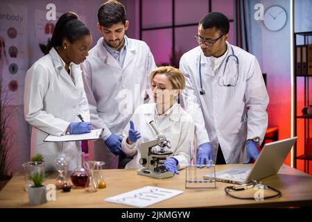 Team of medical research scientists work on a new generation disease cure. They use microscope, test tubes, laptop and writing down analysis results. Laboratory looks busy, modern, night time. Stock Photo