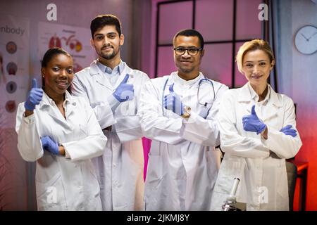 Smiling group of scientists in modern laboratory with male african american leader, mix race team of scientific researchers in lab wearing white coats and stethoscopes, showing thumb up Stock Photo