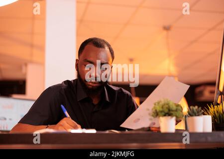 African american employee taking notes about startup contract, analyzing paperwork with business data in office. Working with documents to do research and find executive solutions. Stock Photo