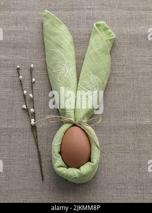 Decorative Bunny Ears from green napkin with egg on natural cotton tablecloth to Celebrate Easter, DIY, flat lay Stock Photo