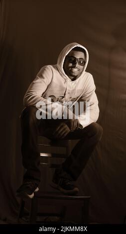 A vertical shot of an African American young male wearing a white hoodie and sitting on a chair Stock Photo