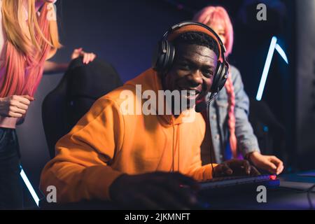 Young black man wearing orange hoodie and headset laughing looking at the camera playing online game supported by two friends. High quality photo Stock Photo