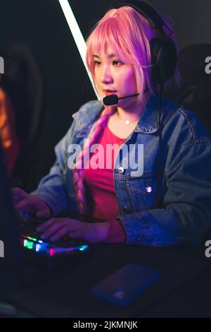 Asian girl with bright pink hair wearing a braid and headset focused playing an online team game using professional gaming setup. Vertical shot. High quality photo Stock Photo