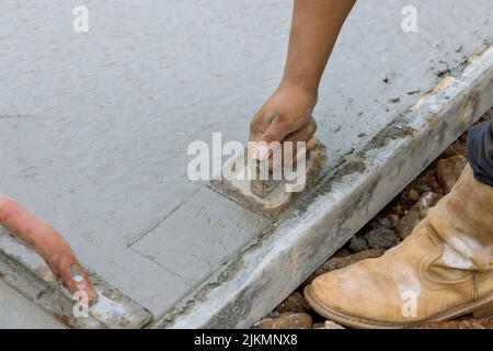 This worker is holding a steel trowel and smoothing out the plastering of a new sidewalk on a freshly laid concrete slab Stock Photo