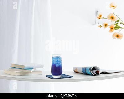 A glass of refreshing blue drink and newspapers on a table with white curtains in the background Stock Photo