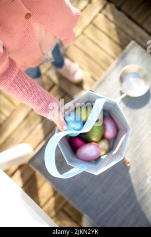 A vertical top view of a children's hand putting a colorful egg in the bag full of egs Stock Photo