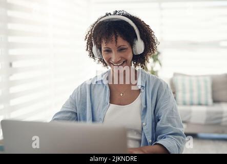 Setting the tone for work. an attractive young woman sitting alone at home and using her laptop while wearing headphones. Stock Photo