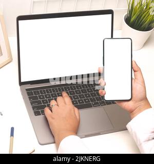 A laptop on the table and a smartphone in a hand with blank screens for copy space Stock Photo