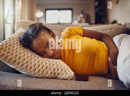 Stomach pain, illness and sickness causing a tired, young and sleepy black woman to sleep, nap and rest on a couch at home. Relaxed, sick or ill Stock Photo