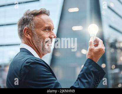 The best tool in a business kit is a bright mind. a mature businessman holding a lightbulb against an urban background. Stock Photo