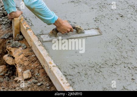 Currently, a worker is holding a steel trowel and smoothing plaster on freshly poured concrete sidewalk Stock Photo