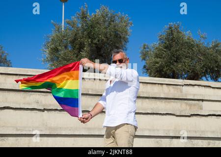 Handsome mature man with beard and grey hair is sitting on the bleachers of an open-air auditorium while holding the gay pride flag in his hands. Conc Stock Photo